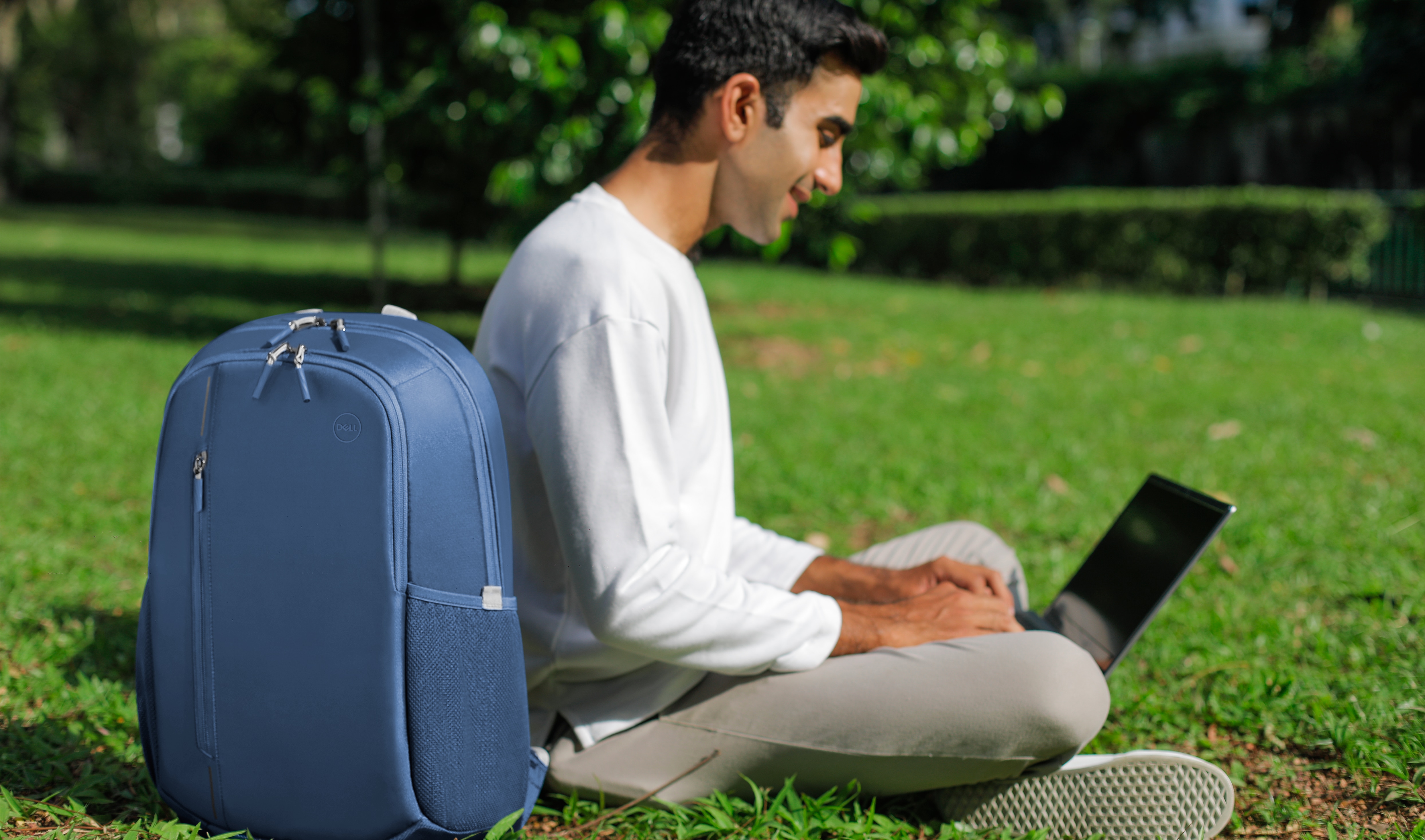 Imagem de um homem sentado na grama com um notebook Dell no colo e uma mochila azul Dell CP4523B ao lado.