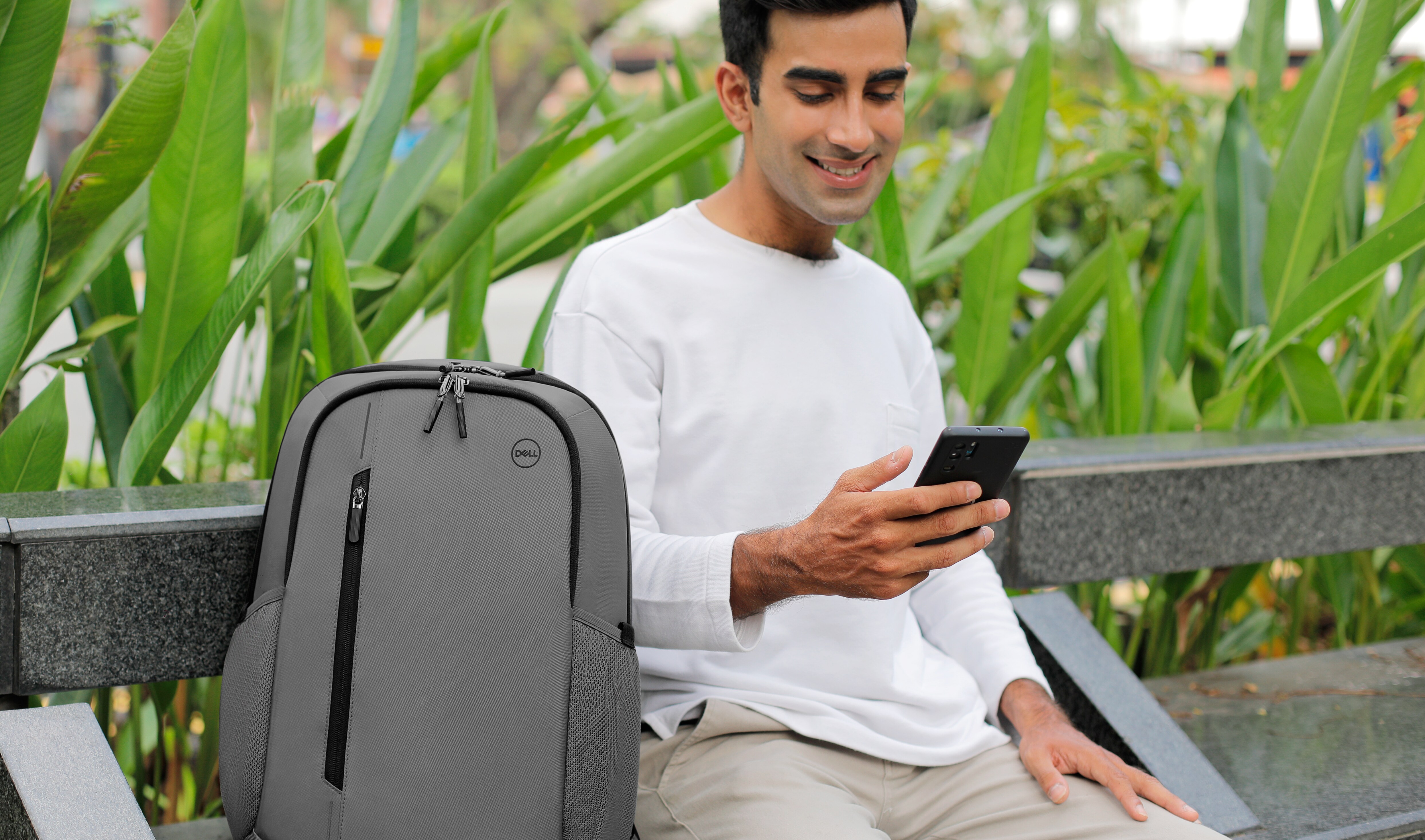 Imagem de um homem sentado em um banco com um celular na mão e uma mochila cinza Dell CP4523G ao lado.