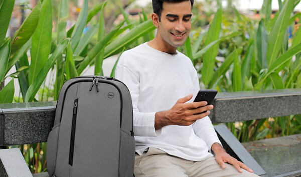 Imagen de un hombre sentado en un banco con un teléfono celular en la mano y una mochila Dell CP4523G gris a un lado.